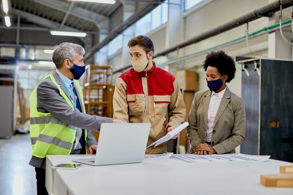Factory manager and workers in masks discussing plans with laptop.