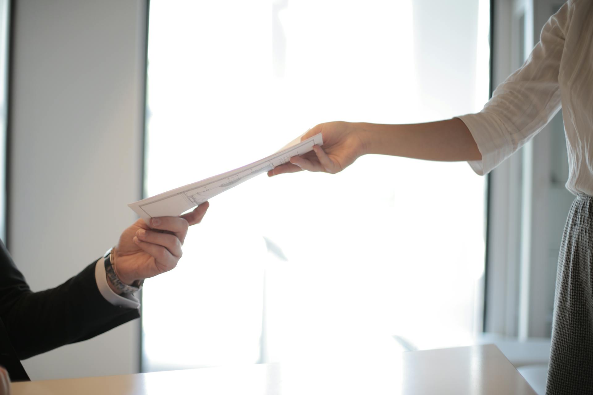 Person handing over documents to another individual across a desk.
